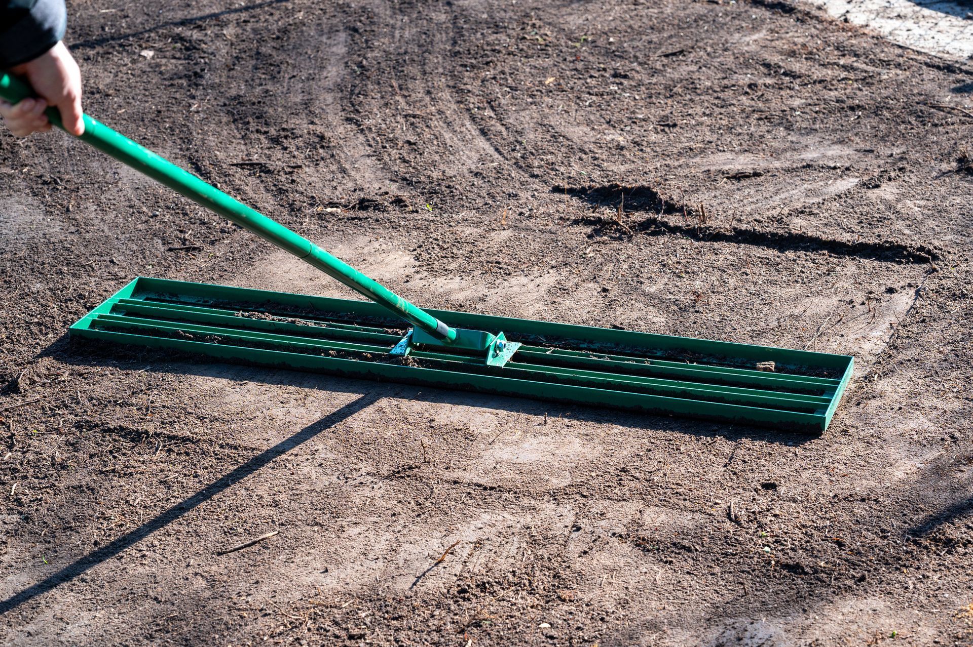 A person uses a long-handled green landscape rake to level dark soil on a patch of ground.