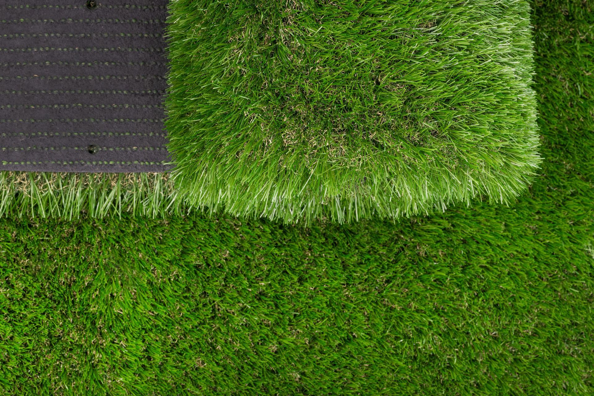 A section of artificial turf being rolled out onto an existing grass surface, showing the black backing and green blades.