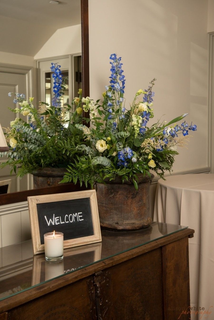Welcome sign with candle on a wooden table beside a blue and white floral arrangement.