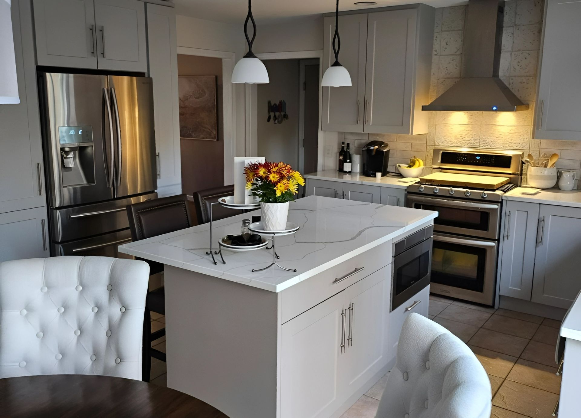 A modern kitchen with grey cabinets, stainless steel appliances, a white quartz island, and two pendant lights overhead.