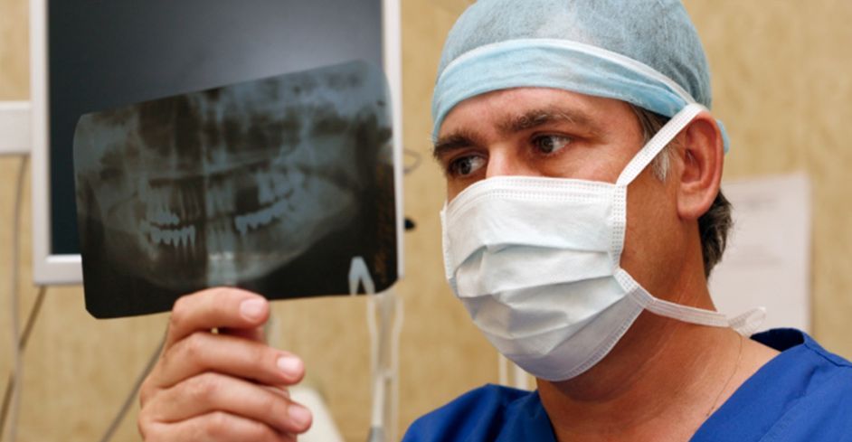 Un dentista con uniforme azul y mascarilla examina una radiografía dental en un entorno médico.