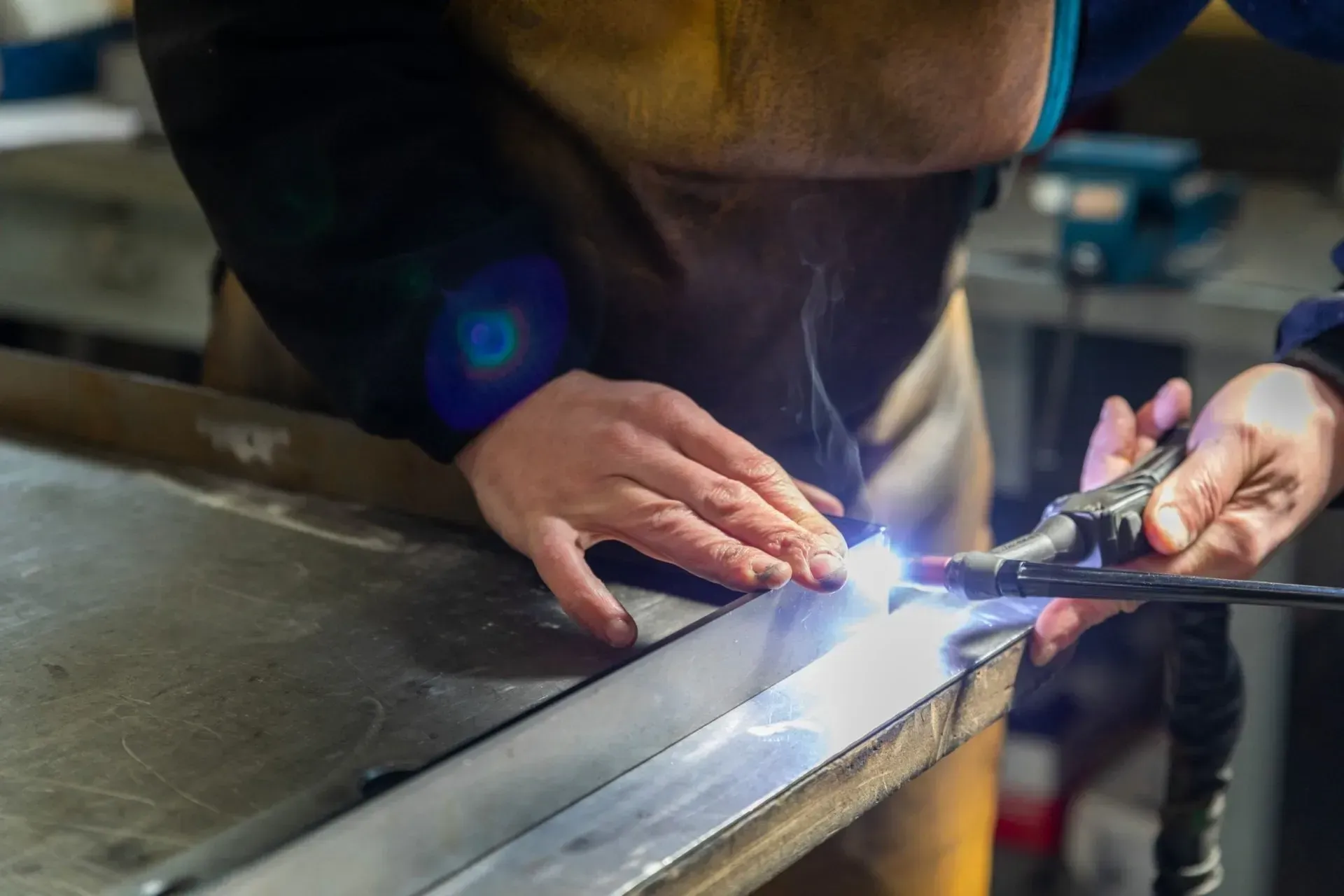 Primo piano delle mani di un operaio che utilizza una torcia per saldatura TIG per unire un pezzo di metallo su un banco da lavoro.