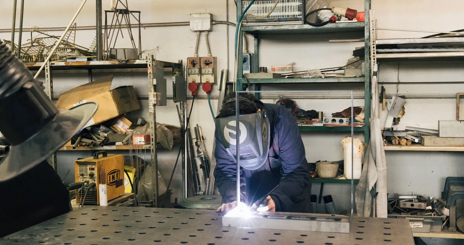 Un operaio con indosso un casco protettivo per saldatura salda un componente metallico su un banco da lavoro perforato in un'officina disordinata.