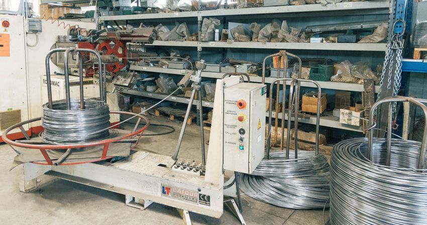 Avvolgitrice industriale per fili metallici con bobine su supporti metallici in un'officina, con scaffalature di stoccaggio sullo sfondo.