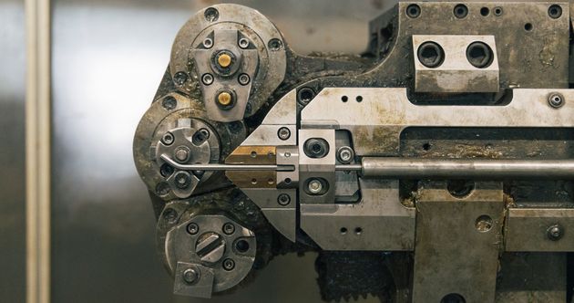 Primo piano di una piegatrice industriale per fili metallici che modella una barra di metallo con teste meccaniche di precisione.