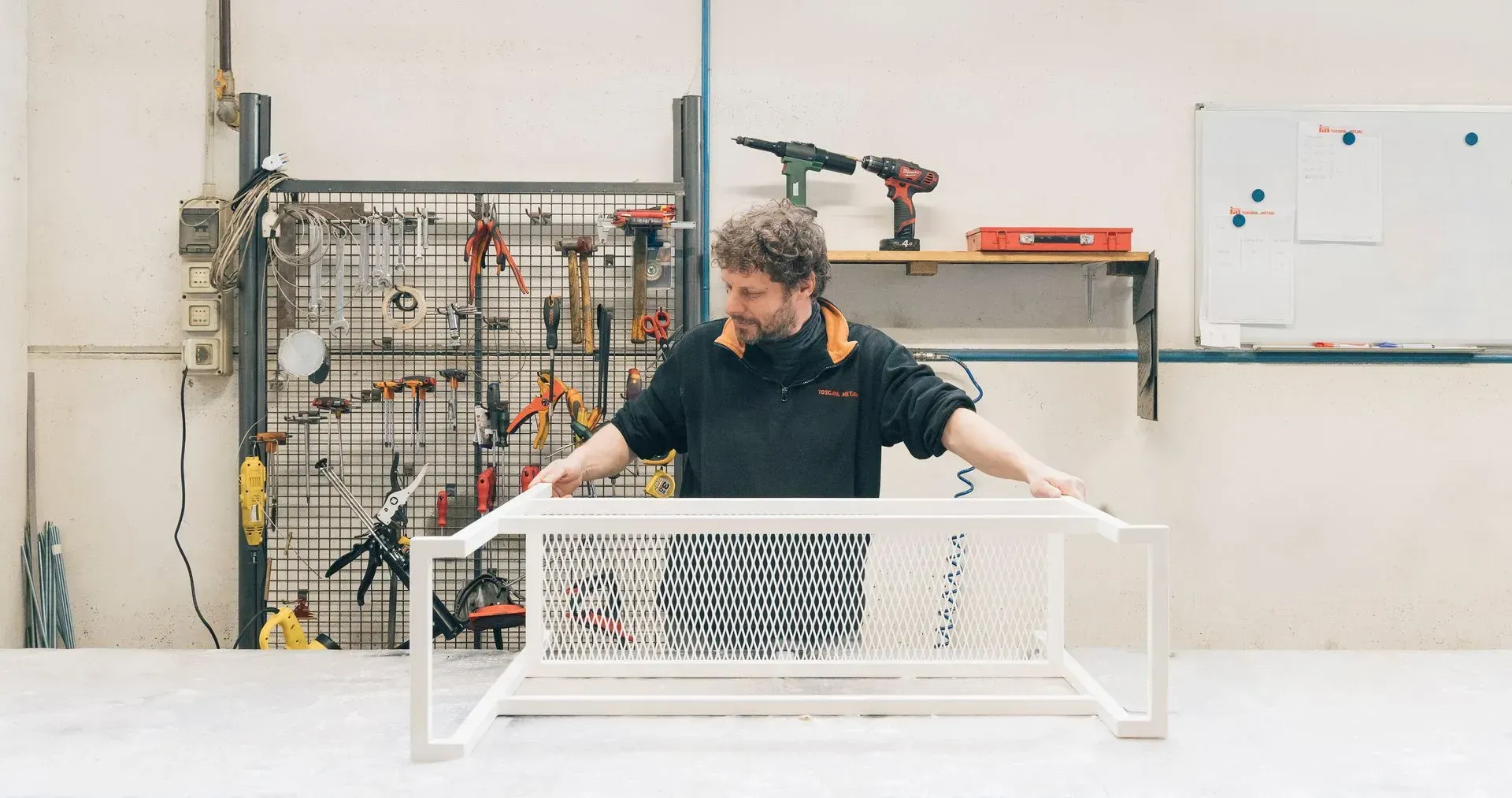 In un'officina, una persona assembla su un tavolo la struttura di un mobile in metallo bianco con motivo a griglia.