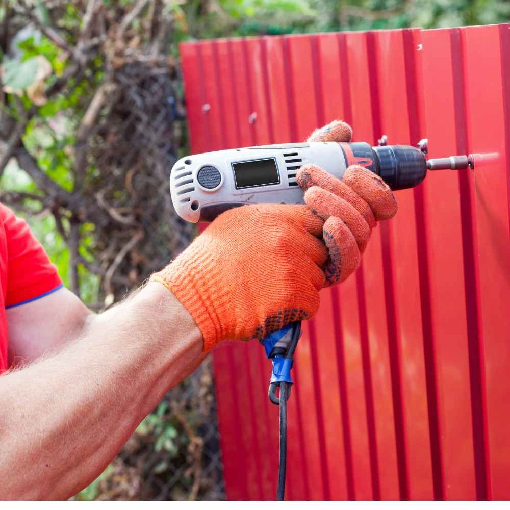 A man is holding a drill in his hand in front of a red fence.