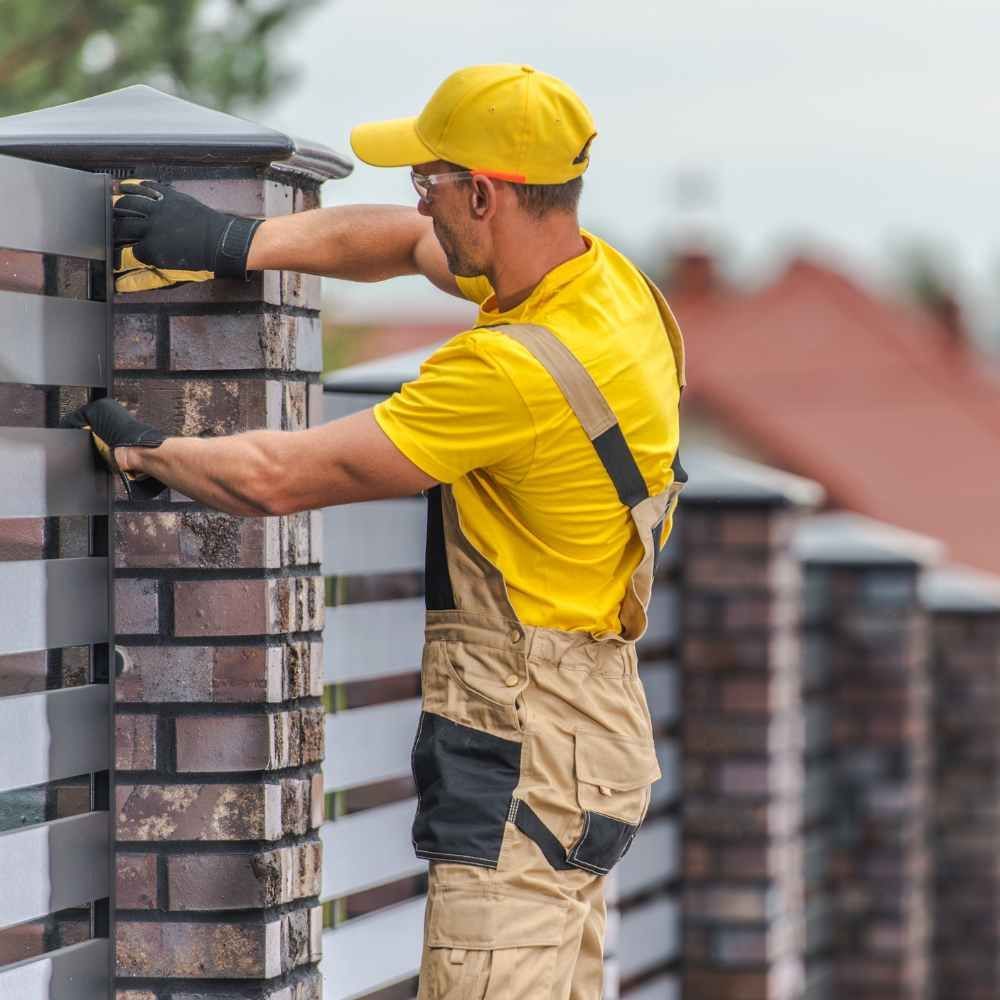 A man in a yellow shirt is working on a brick fence.