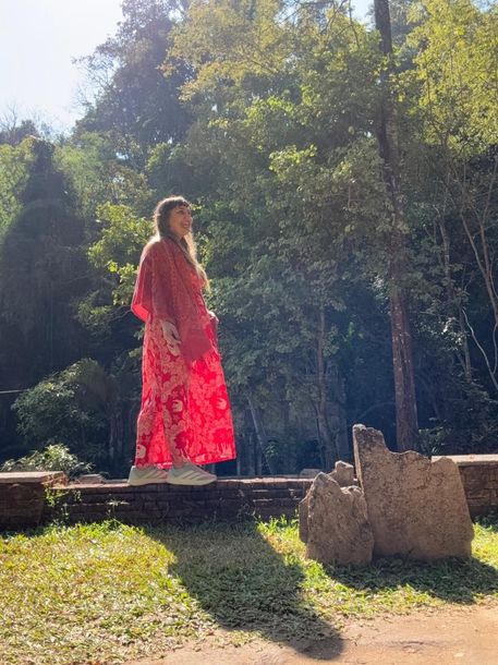 Una persona con un vestido rojo brillante está de pie sobre una plataforma de madera en un claro soleado del bosque.