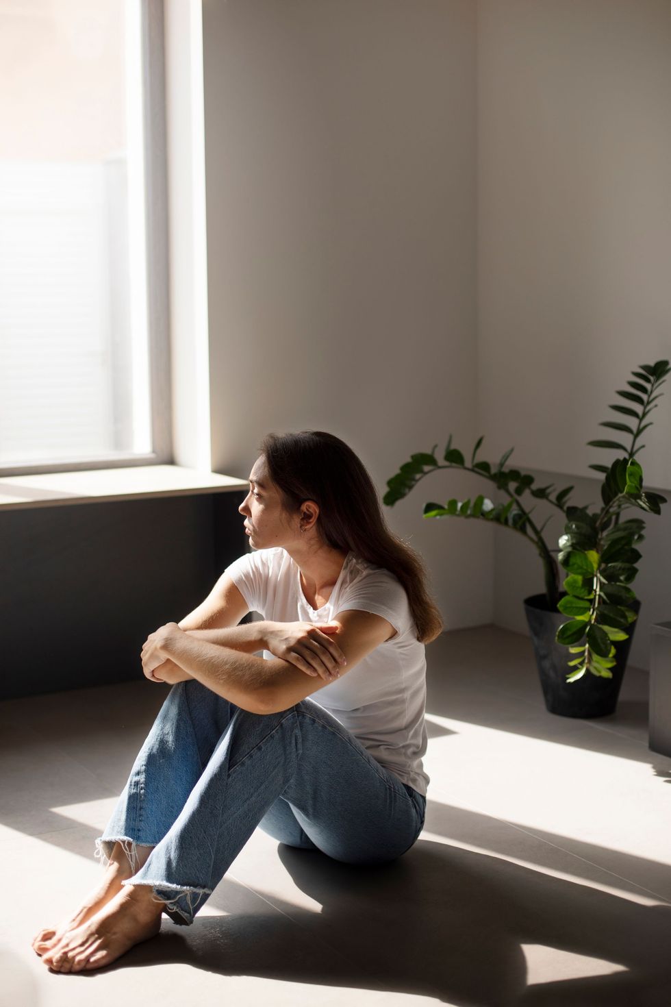 Una mujer sentada en el suelo junto a una ventana soleada, abrazando sus rodillas junto a una maceta.