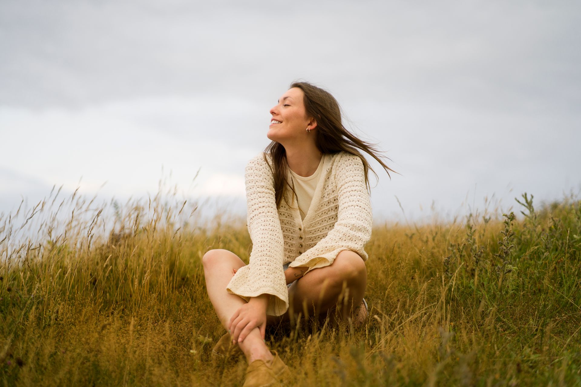 Una persona sentada en la hierba alta, sonriendo hacia un cielo nublado, con un suéter ligero.