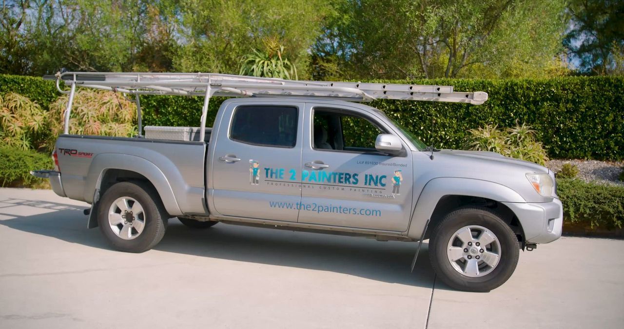 Silver pickup truck with ladders on the roof rack, parked in a driveway. The truck has company signage.