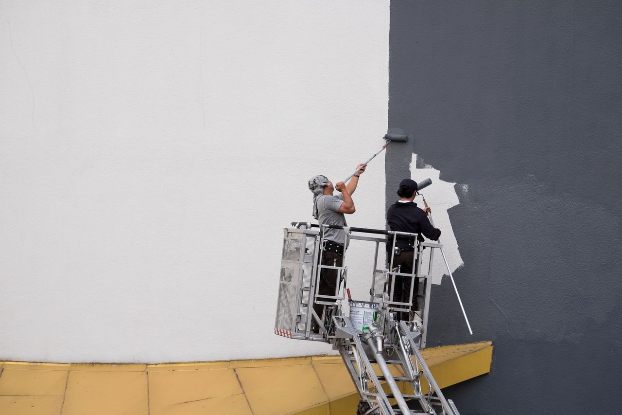 Two people on a lift paint a building wall, one using a roller, switching from white to gray.