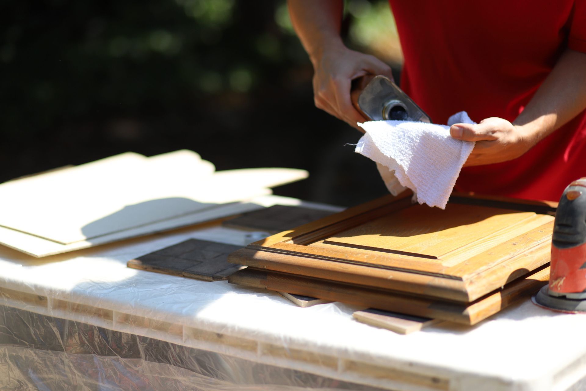 Person wiping wood cabinet door with a cloth outside, other cabinet pieces on the table.