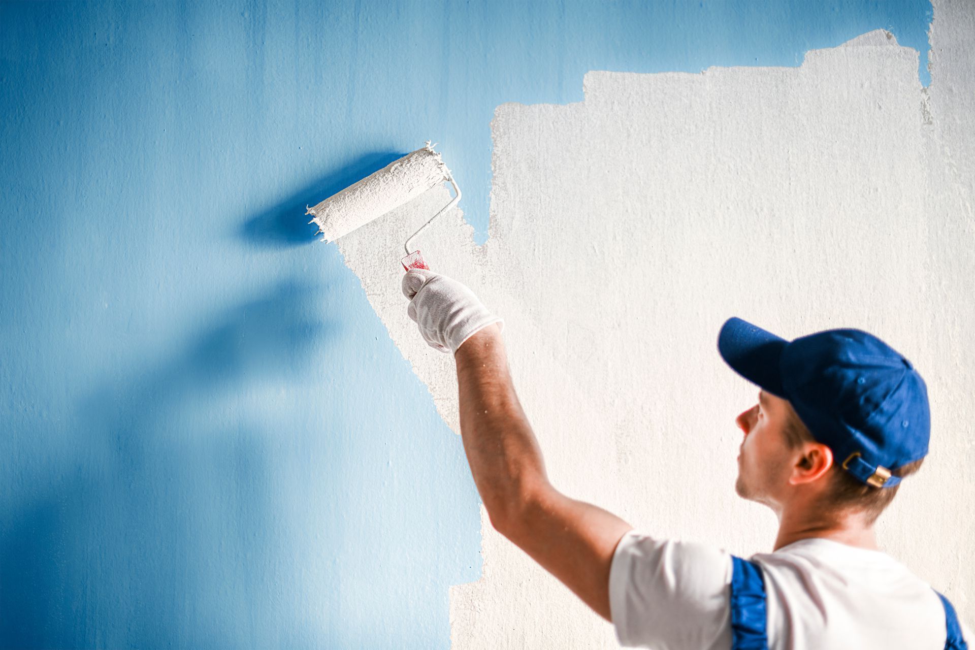 Person painting a blue wall white with a roller; wearing a blue cap and overalls.