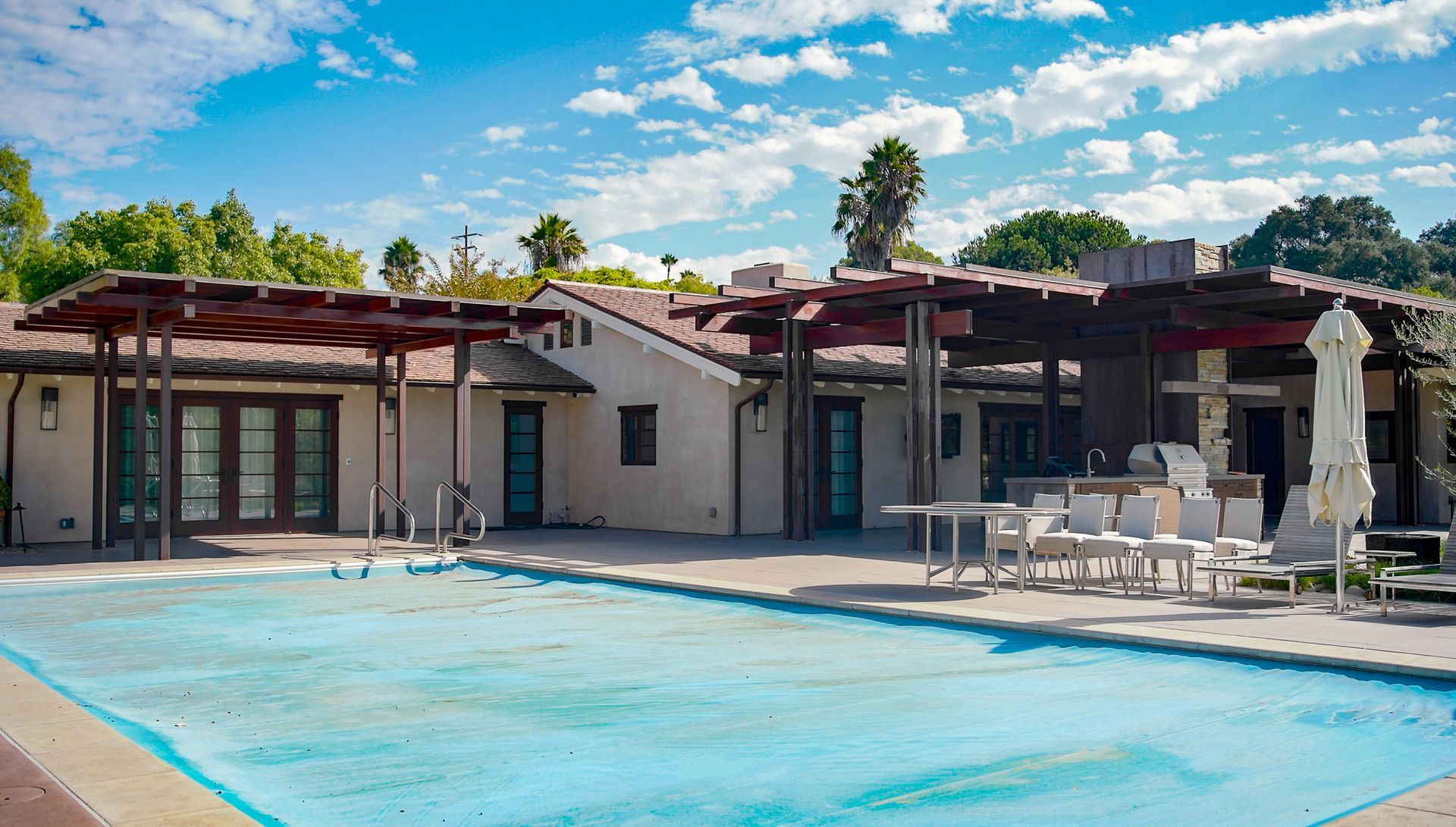 A swimming pool in front of a tan building with a shaded patio area, against a blue sky.