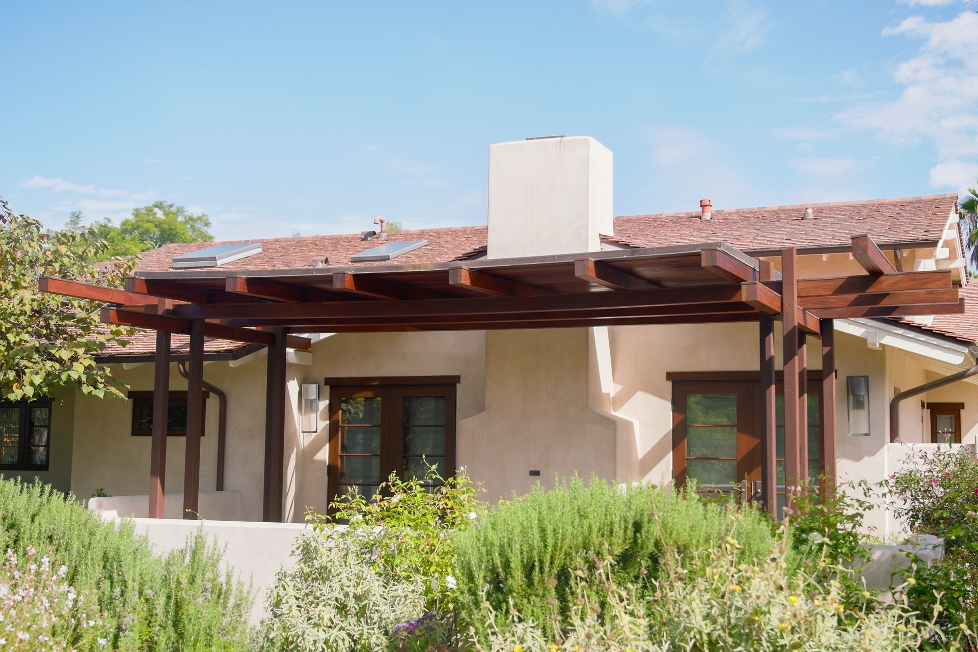 Brown wooden pergola over a stucco house with a red tile roof and surrounding greenery.
