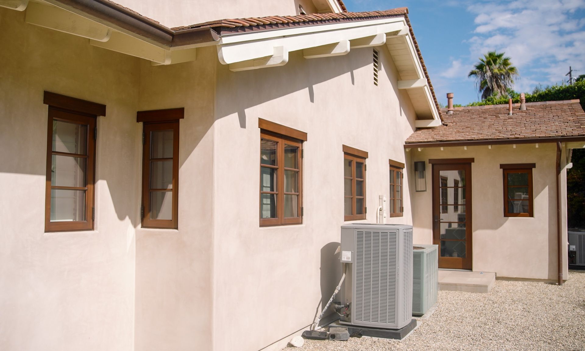 Beige stucco house with brown-framed windows, an air conditioning unit, and a door on a sunny day.