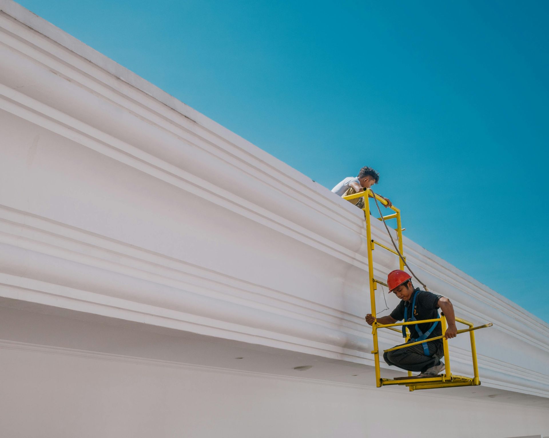 Two workers on a yellow lift, repairing a white building against a blue sky.