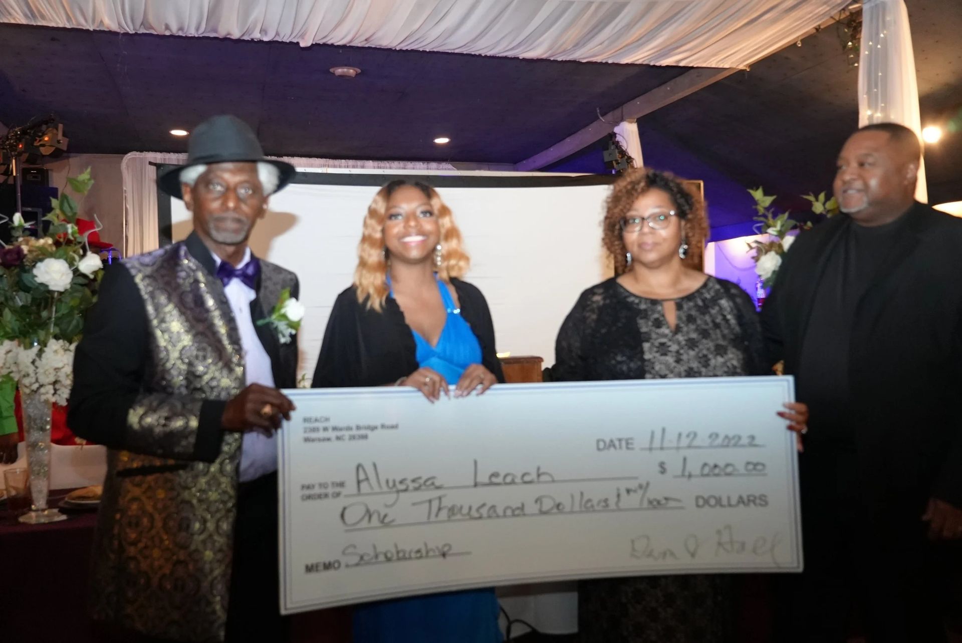 Four people holding a large check. Alyssa Leach receives $2,000. Formal event setting with flowers, a screen, and decorations.