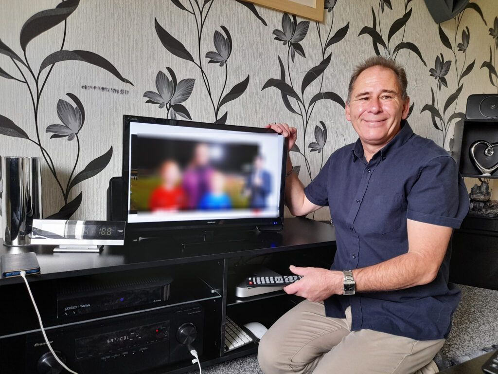 A man is sitting in front of a television holding a screen celebrating a milestone.