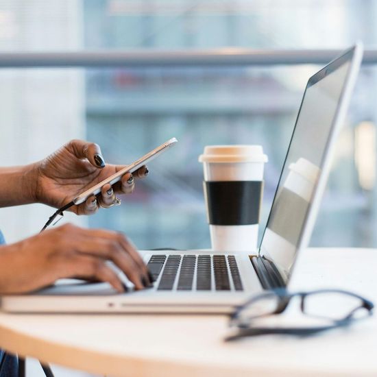 A person is typing on a laptop while holding a cell phone.