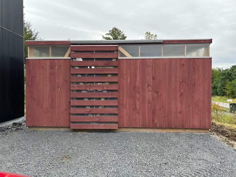 A red wooden shed is sitting on top of a gravel lot next to a black building.