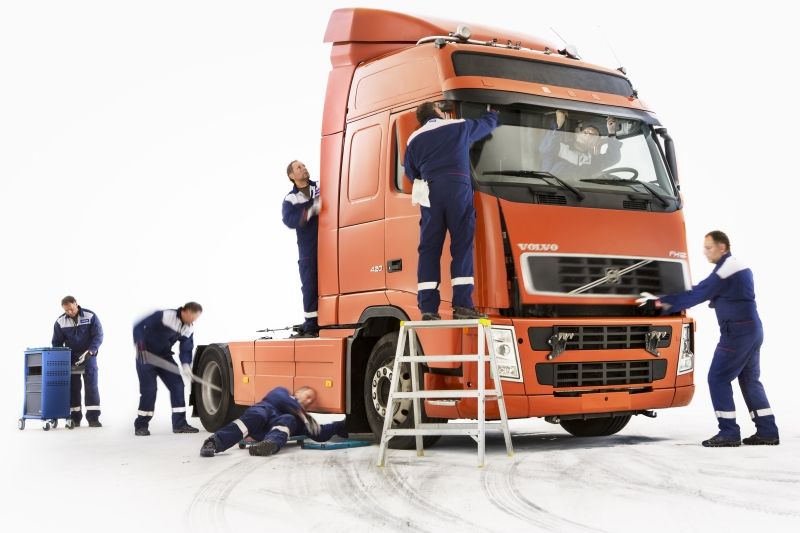 Mecánicos con uniformes azules trabajando en un camión semirremolque naranja en un estudio blanco.