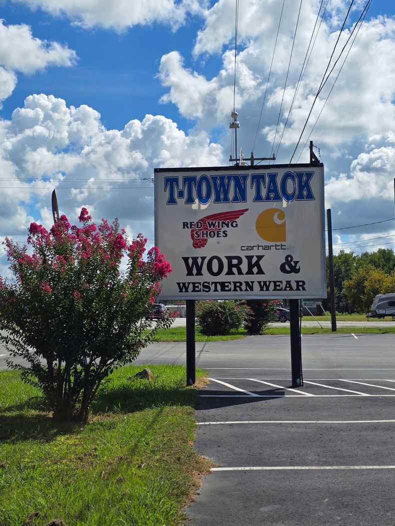 Sign for T-Town Tack shop, with a red flowering bush in front and a partly cloudy blue sky above.