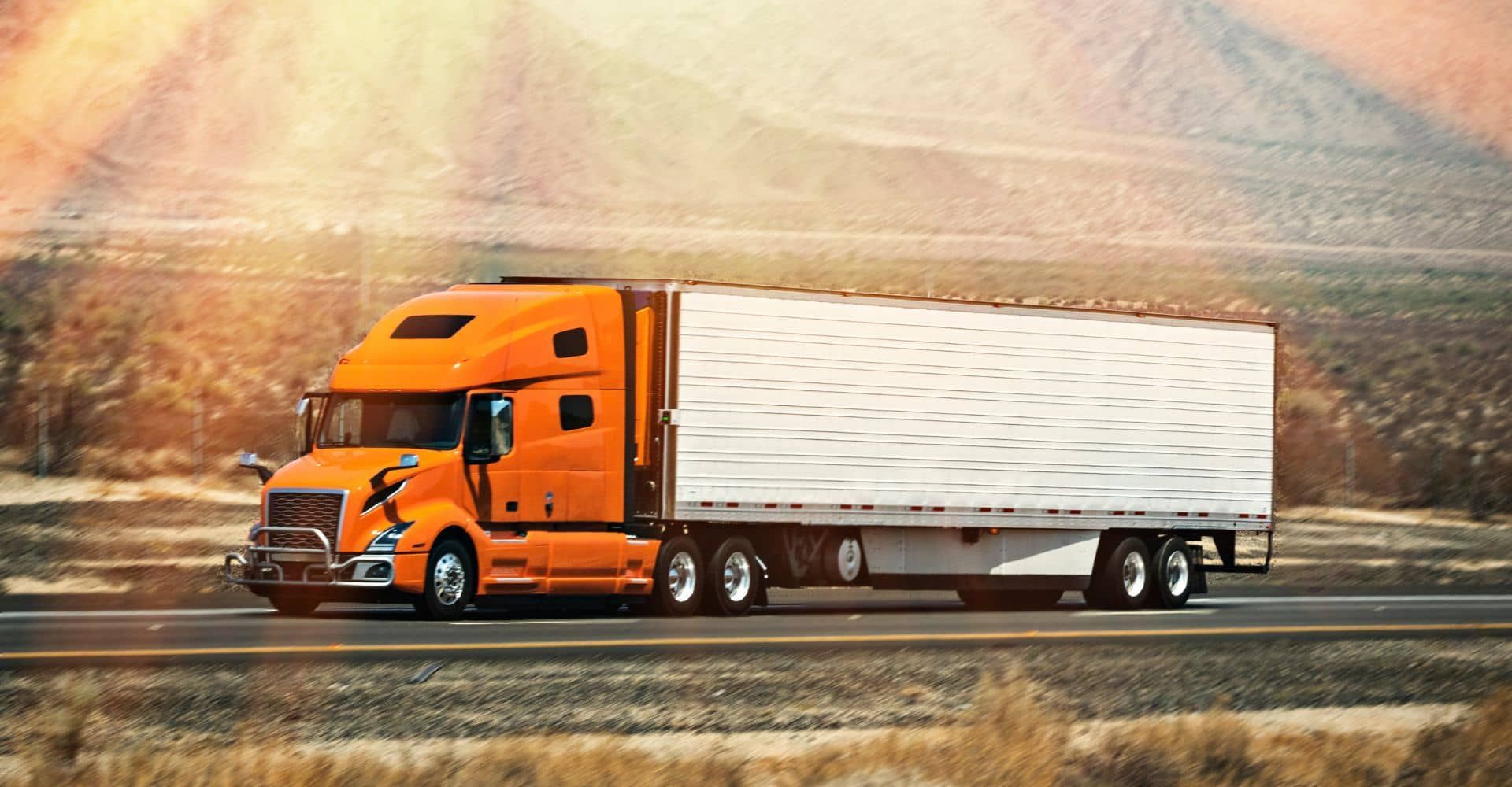 An orange semi truck is driving down a highway in the desert.