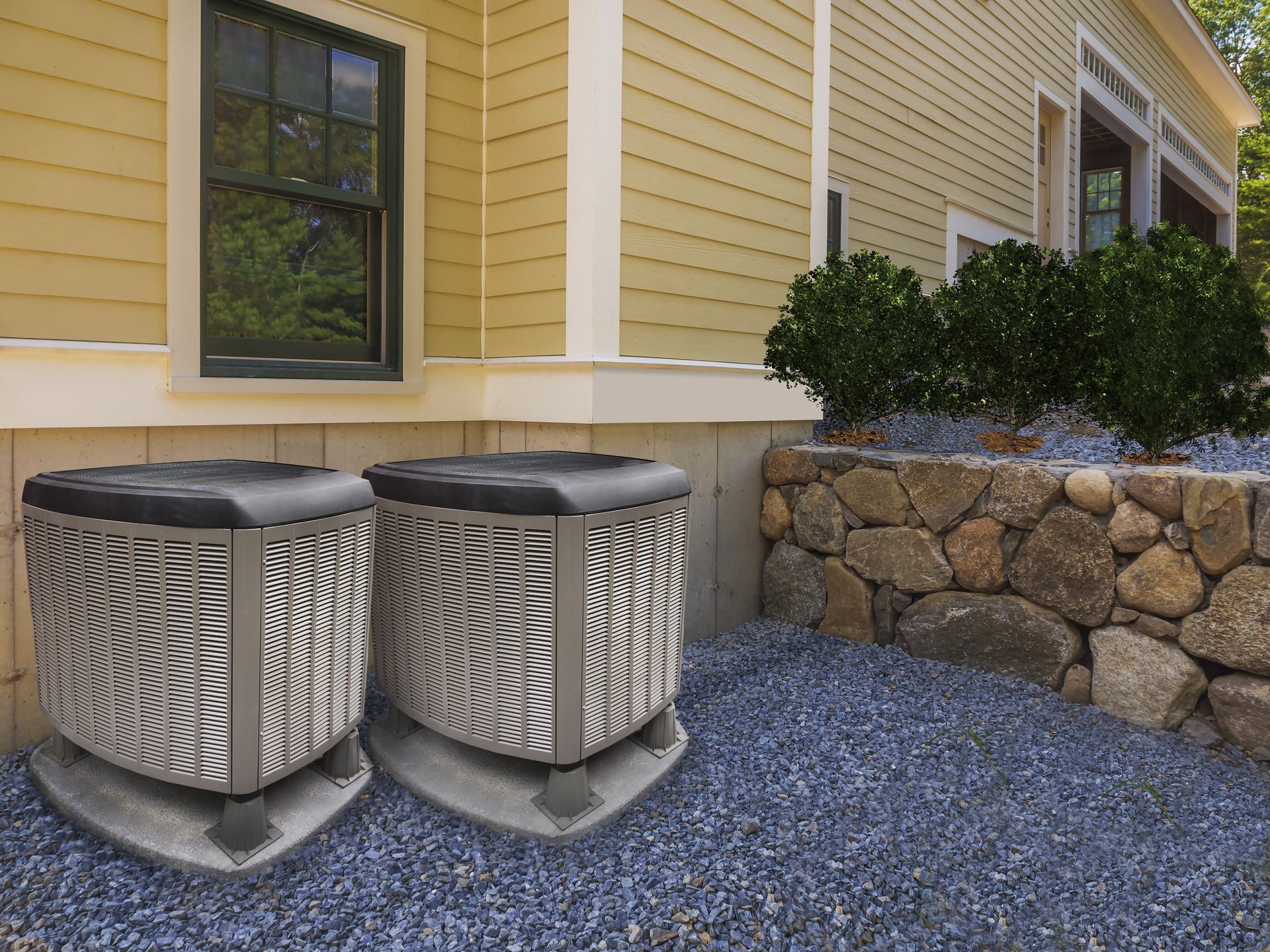 Two air conditioning units on concrete pads next to a building with a rock wall in the background.