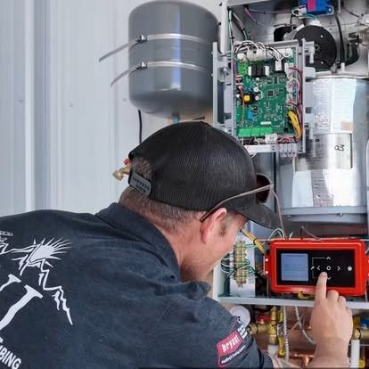 A crane lifts an HVAC unit over a flatbed truck in a parking lot. A building and D.N.I. Heating and Cooling van are in view.