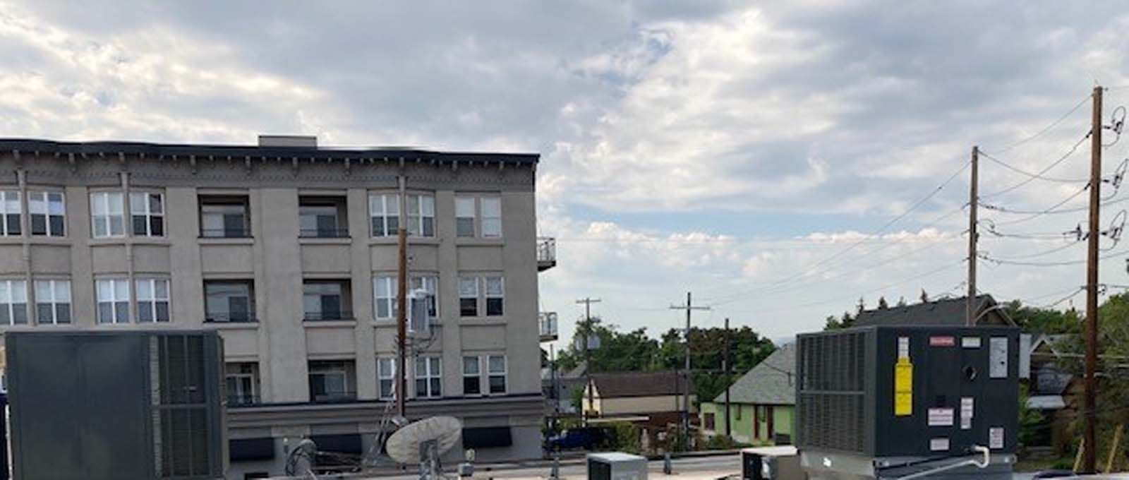 View from a rooftop of buildings and power lines against a cloudy sky. A multi-story building and several boxy structures are in view.
