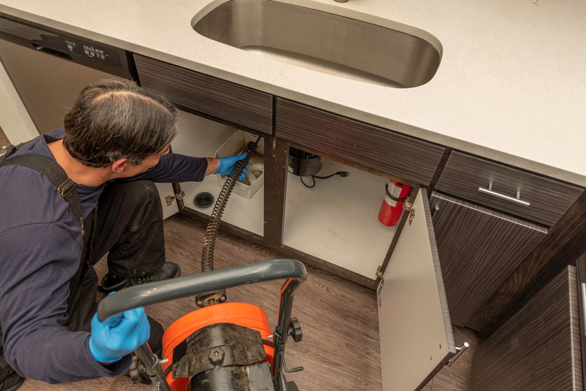 Plumber using a drain snake under a kitchen sink. Orange and black tool, blue gloves, white countertop.