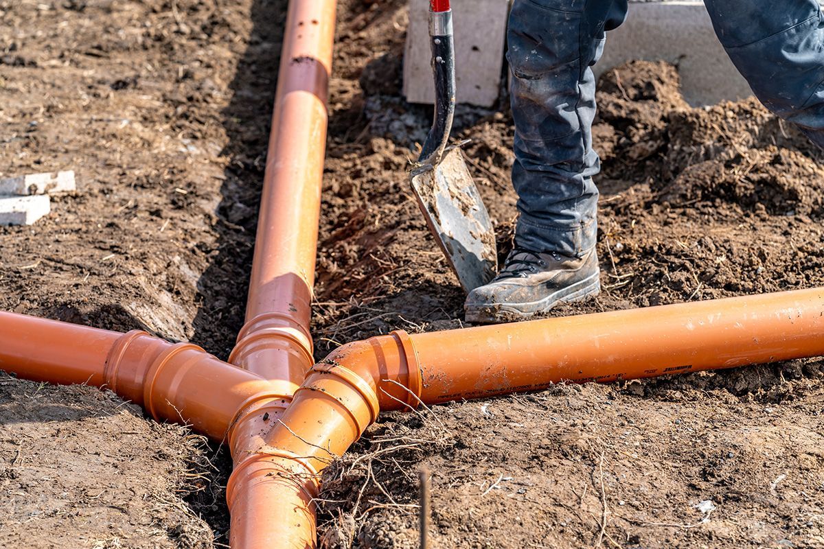 Worker installing orange plastic pipes in a trench, using a shovel in dirt.