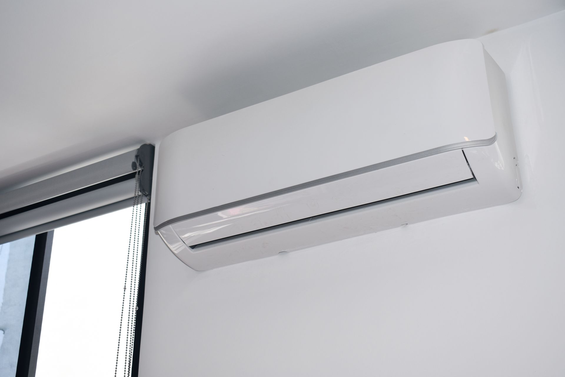 White wall-mounted air conditioning unit near a window with a closed blind.