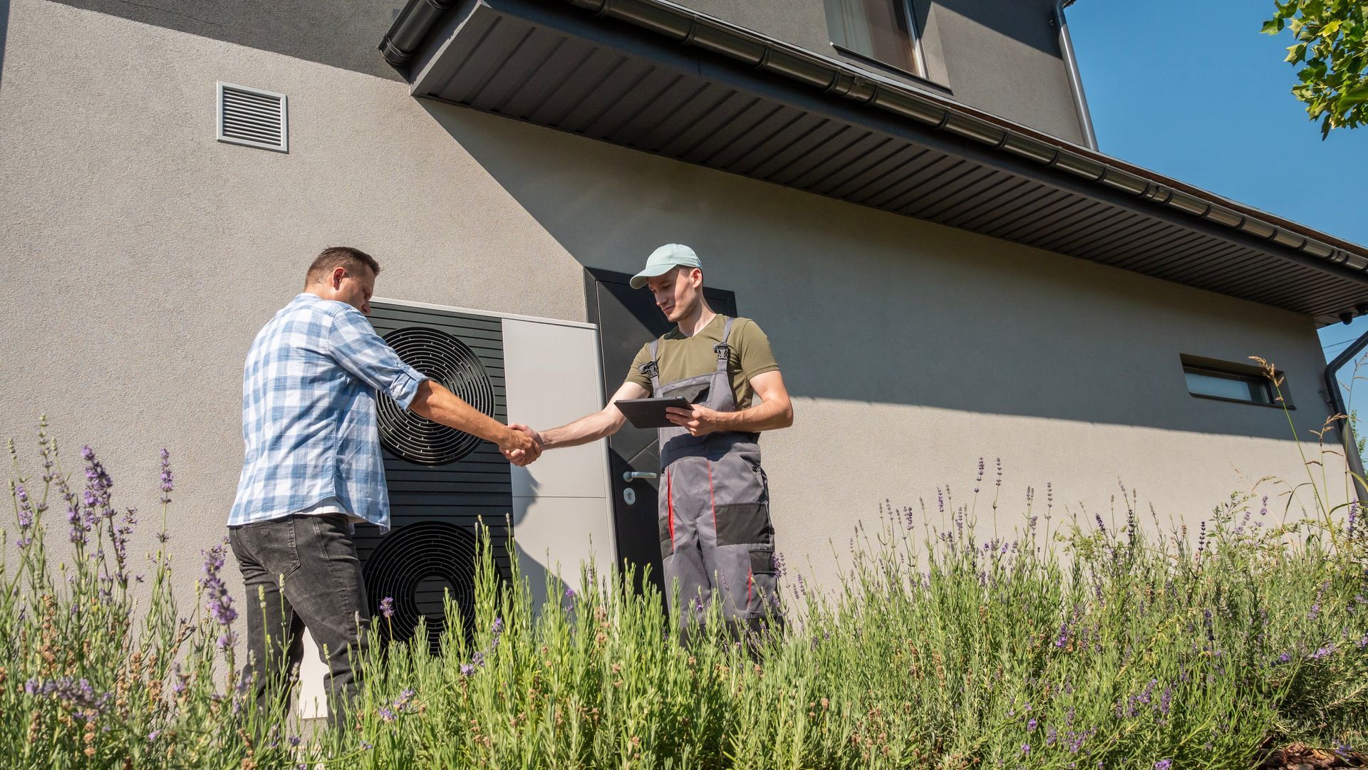 Man shakes hands with a person holding a tablet, standing in front of a house entrance with bushes.