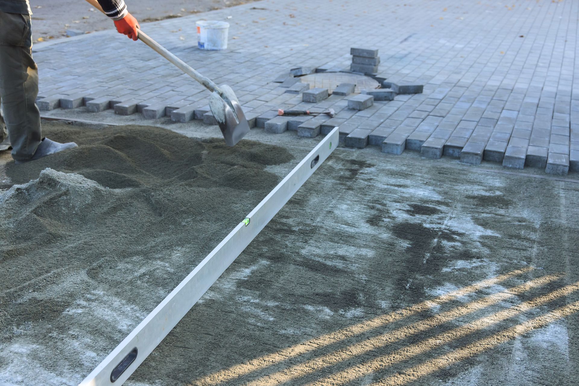 Person shoveling sand, leveling ground for laying gray brick pavers in an outdoor setting.