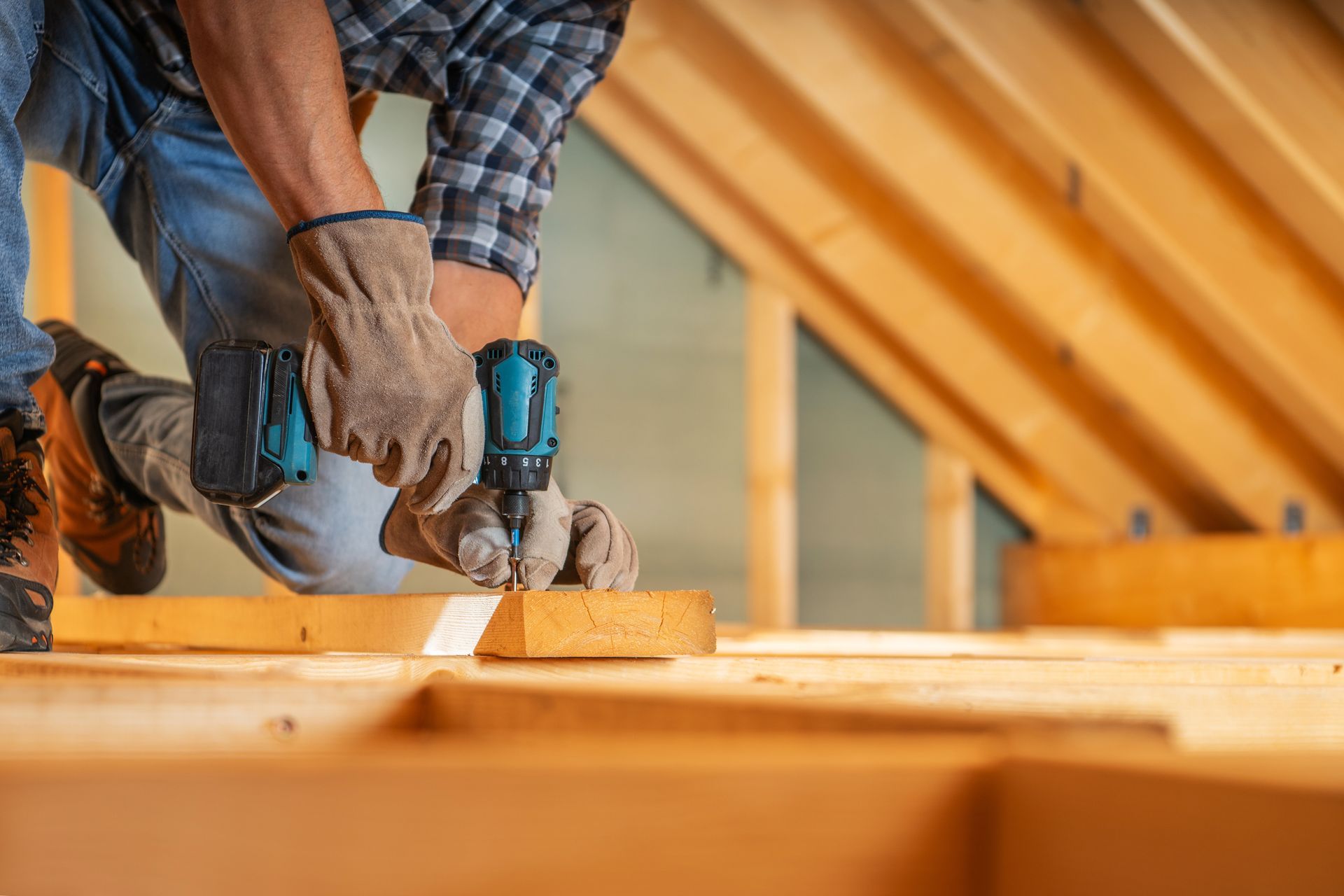 A person wearing work gloves uses a power drill on a wooden beam in an unfinished attic space.