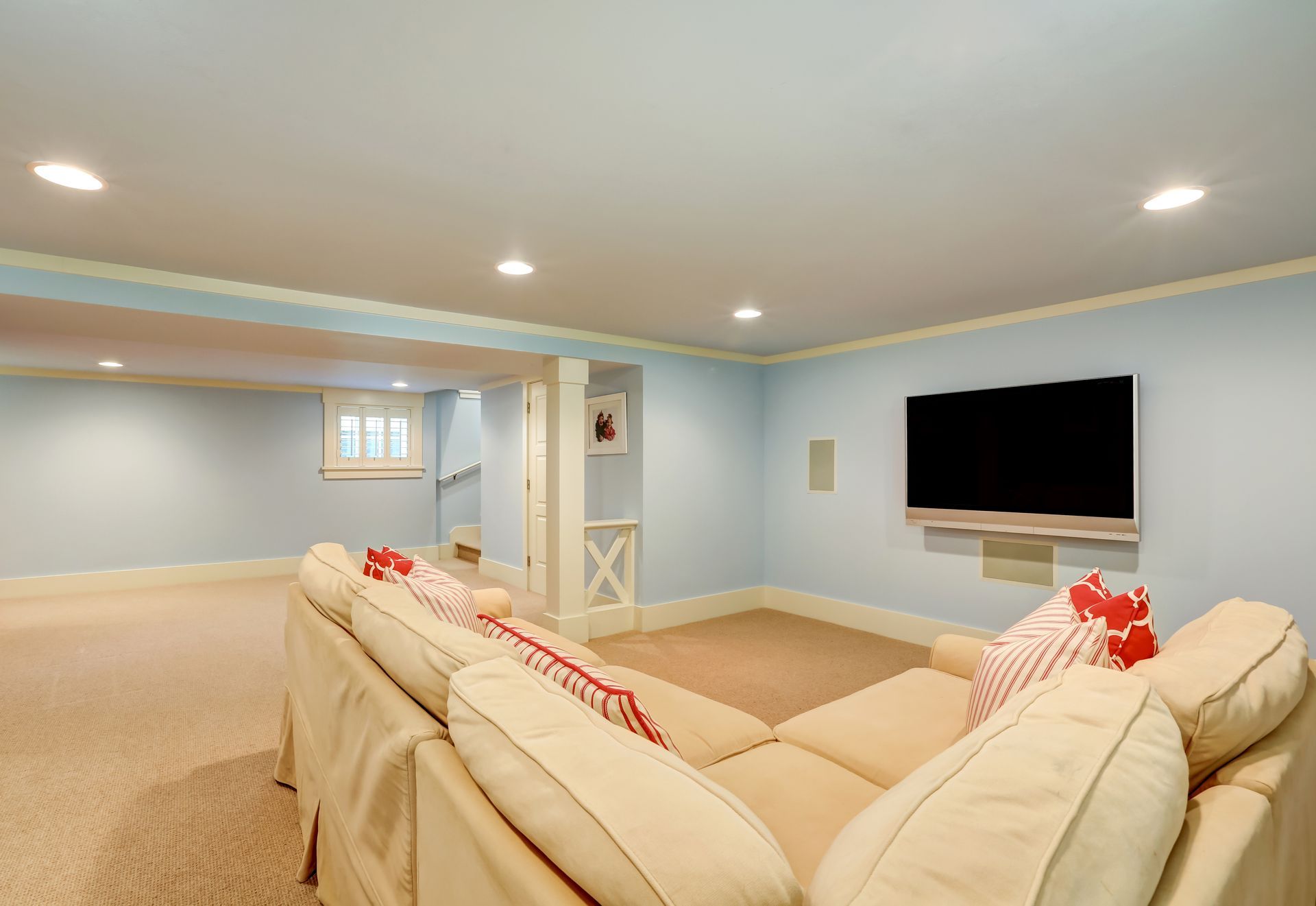 A light-blue finished basement with cream carpeting, a beige sectional sofa, and a television mounted on the wall.