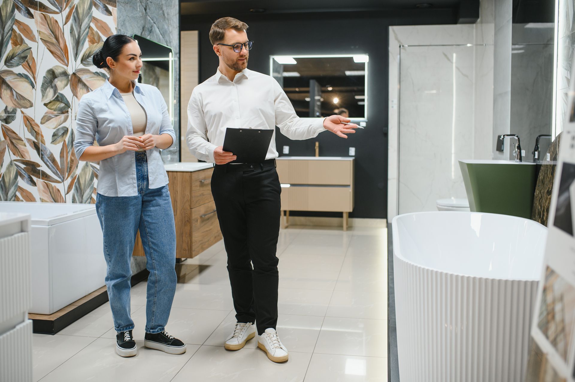 A sales representative holding a clipboard gestures toward a bathtub in a showroom while consulting with a customer.