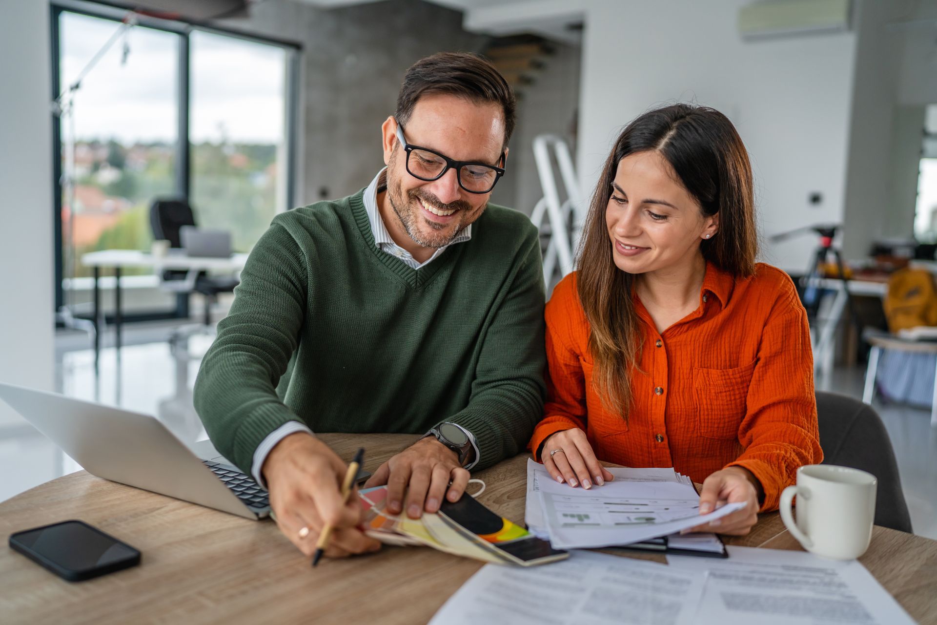 A smiling pair sits at a wooden table in an office, reviewing documents and color samples with a laptop nearby.