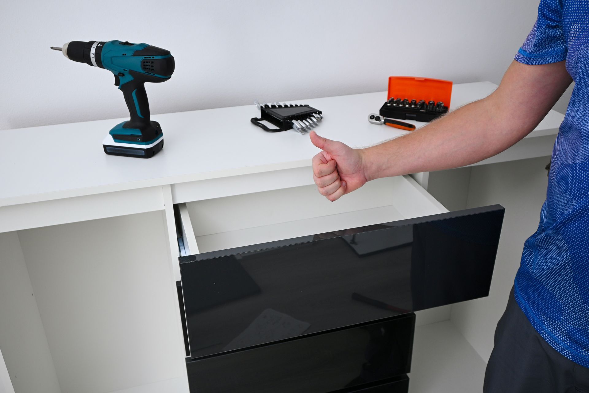 A person giving a thumbs-up next to an assembled white dresser with glossy black drawers and a power drill on top.