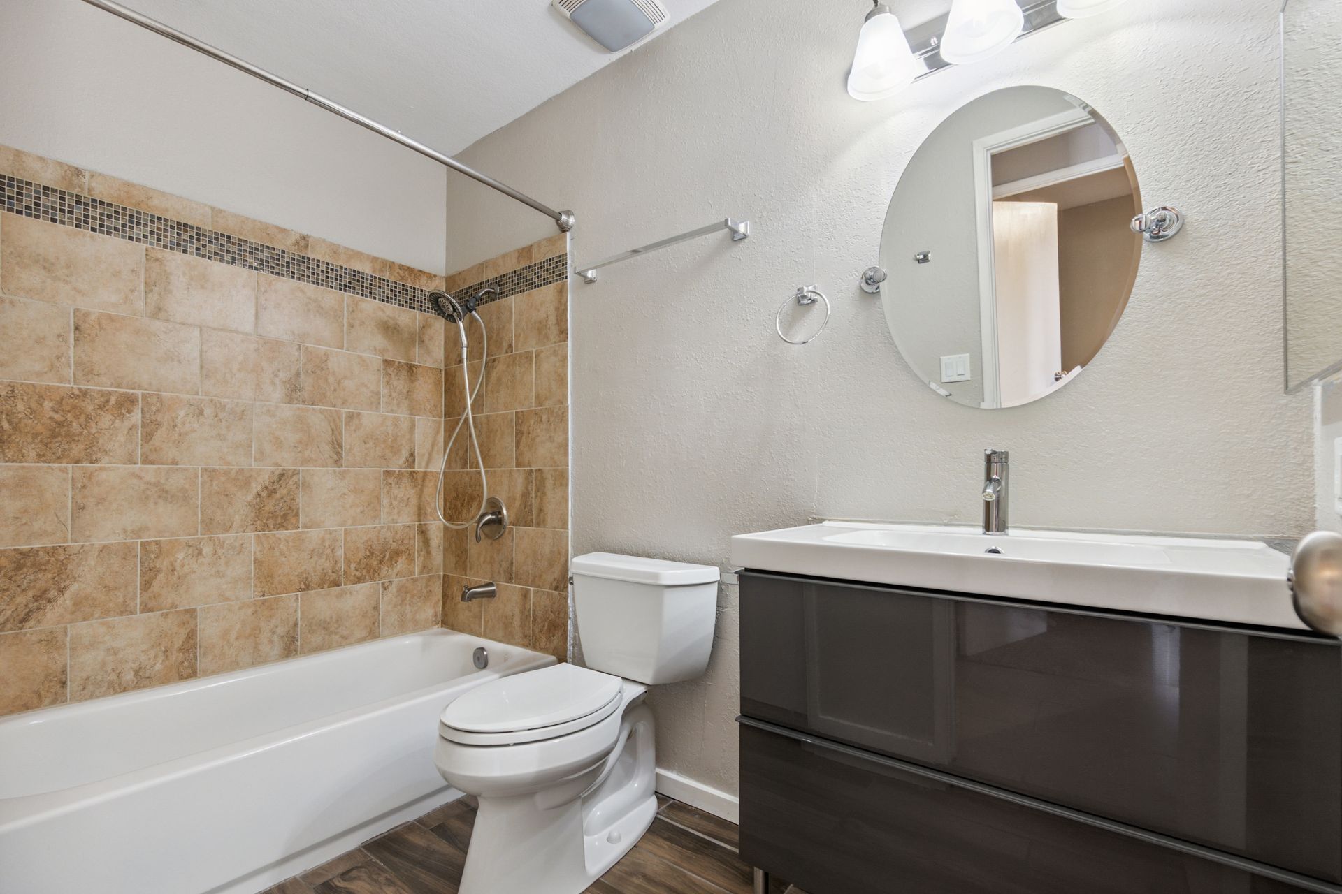 A bathroom with a beige tiled shower, white toilet, and a dark brown vanity with a circular mirror and light fixture.