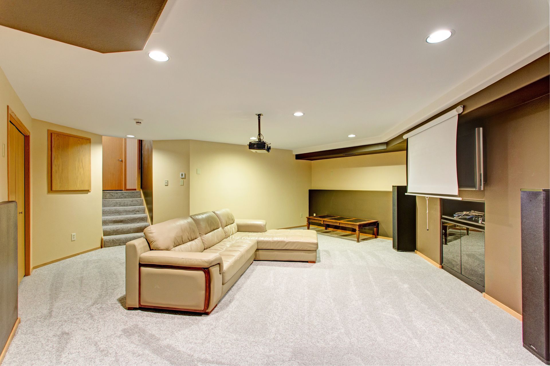 A basement home theater with a beige sectional sofa, projector, retractable screen, and gray carpet.