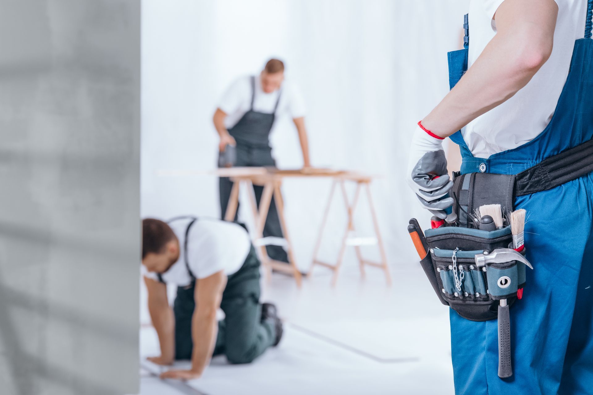 Three construction workers in blue overalls working on an interior renovation, one wearing a tool belt in the foreground.
