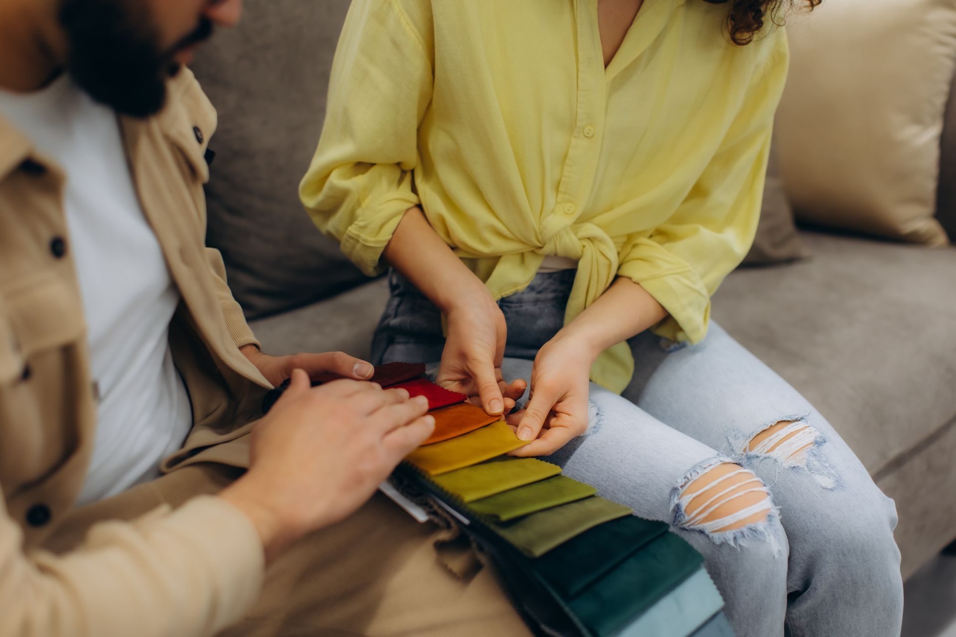 Two people sit on a gray couch, examining and comparing a fabric color swatch book in a well-lit indoor setting.