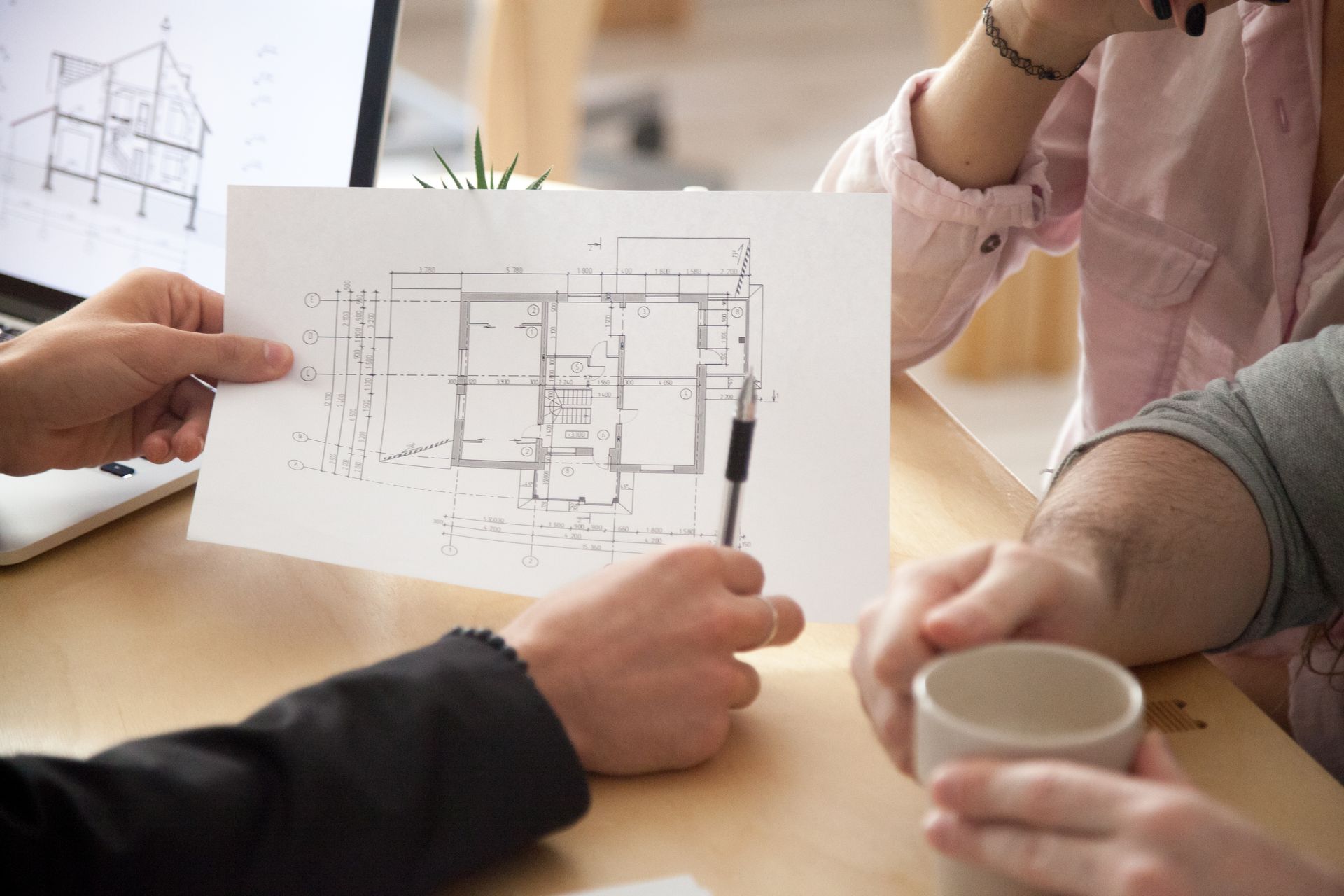 Three people gathered around a table to review a architectural floor plan on paper and a laptop screen.