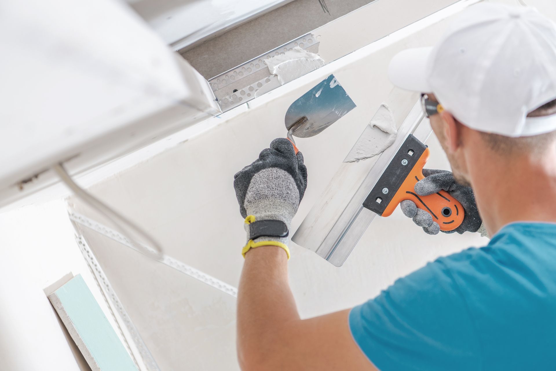 A person wearing gloves and a white cap uses a taping knife and trowel to apply joint compound to a drywall seam.