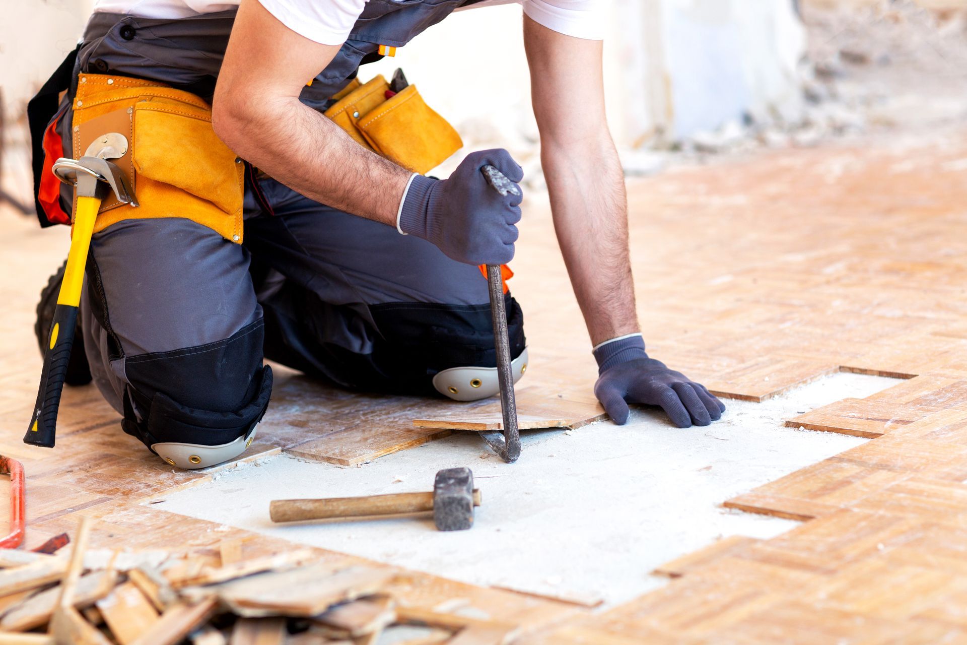 A construction worker in work clothes and gloves uses a pry bar to remove wooden floor tiles.